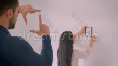 白种人黑发女孩在白墙上选择相框照片的位置，而她的男朋友则用相框造型